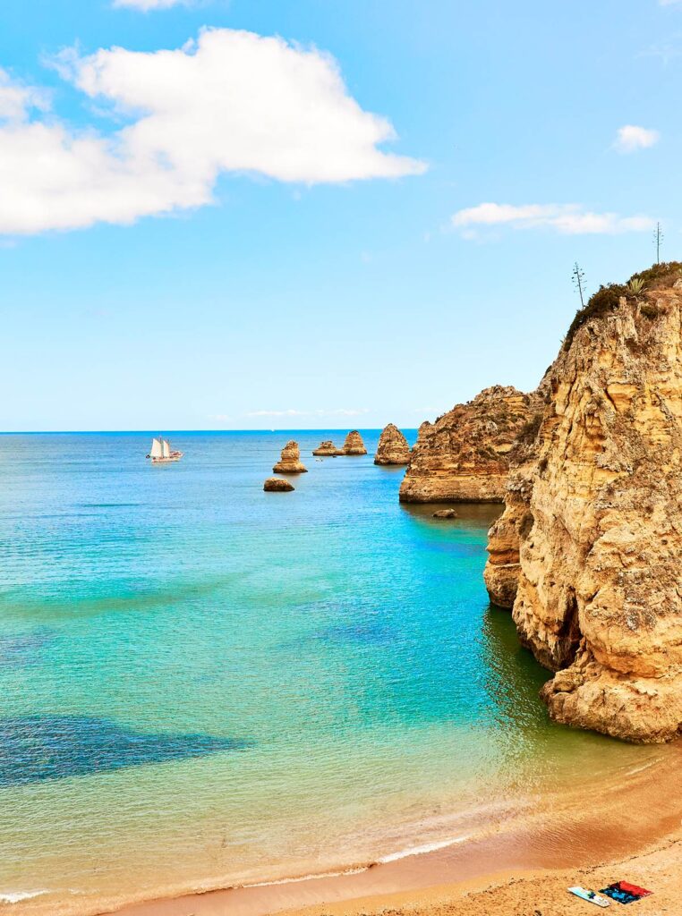Scenic coastal view with turquoise water, rugged cliffs, and a sailing boat under a bright blue sky.
