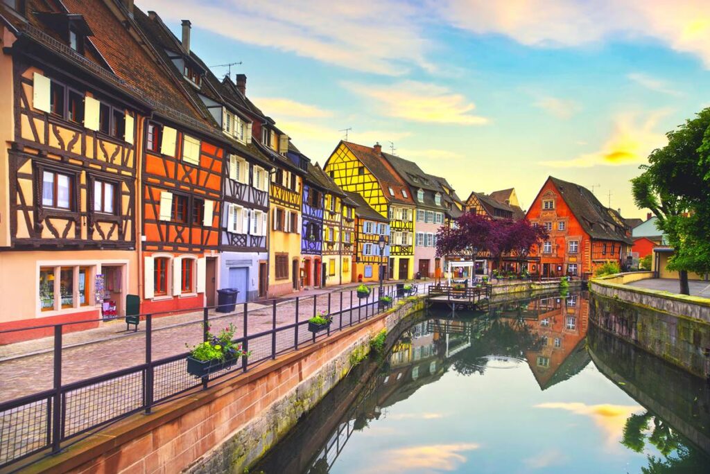 Colorful half-timbered houses along a canal in Colmar, France, reflecting in water under a picturesque sky.