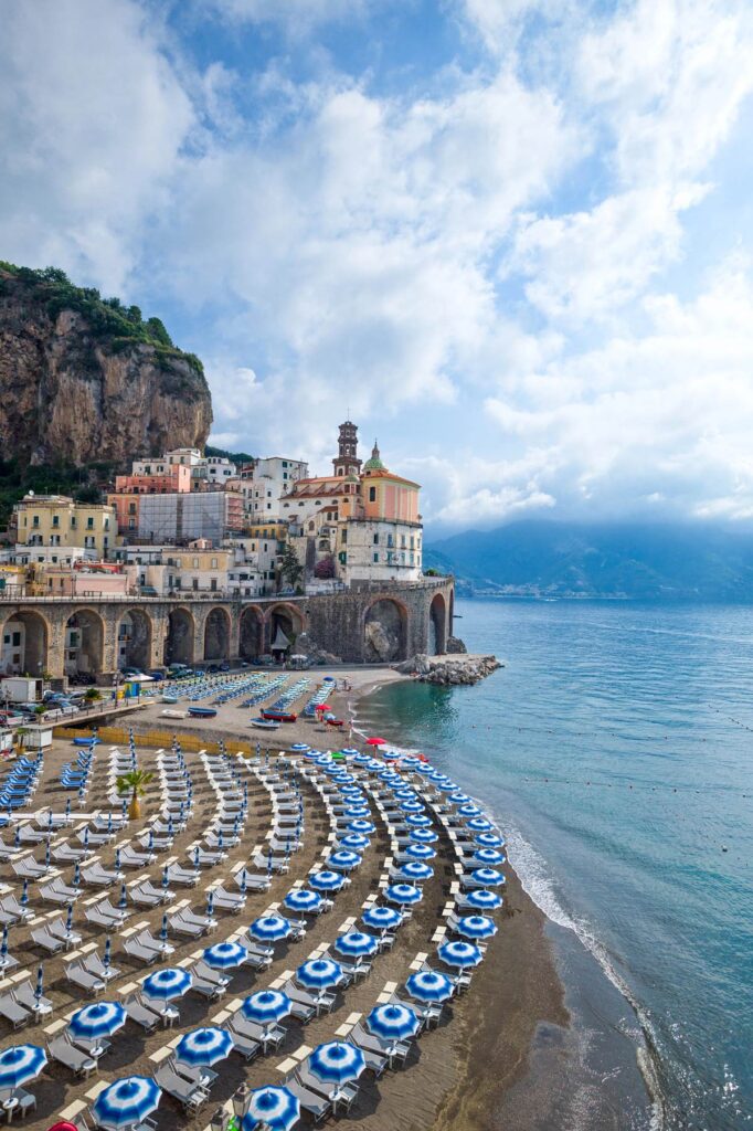 Coastal town with colorful buildings and beach umbrellas, surrounded by cliffs and ocean under a cloudy sky.