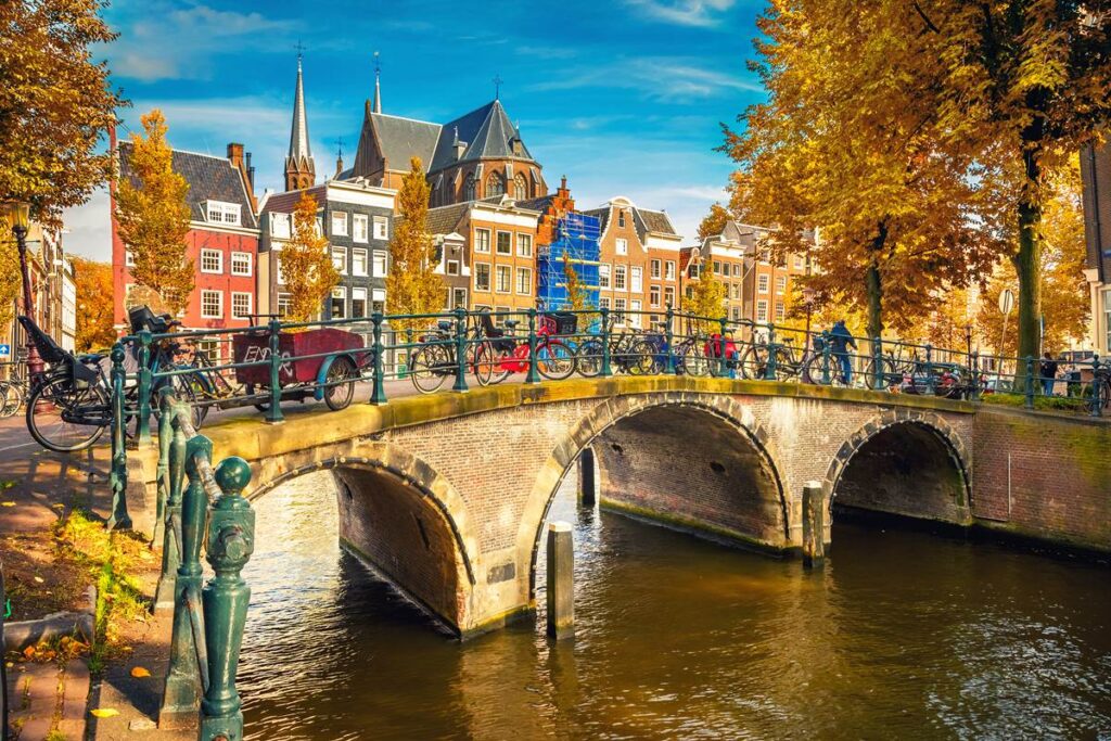 Scenic Amsterdam canal bridge with bicycles, autumn trees, and historic buildings under a clear blue sky.