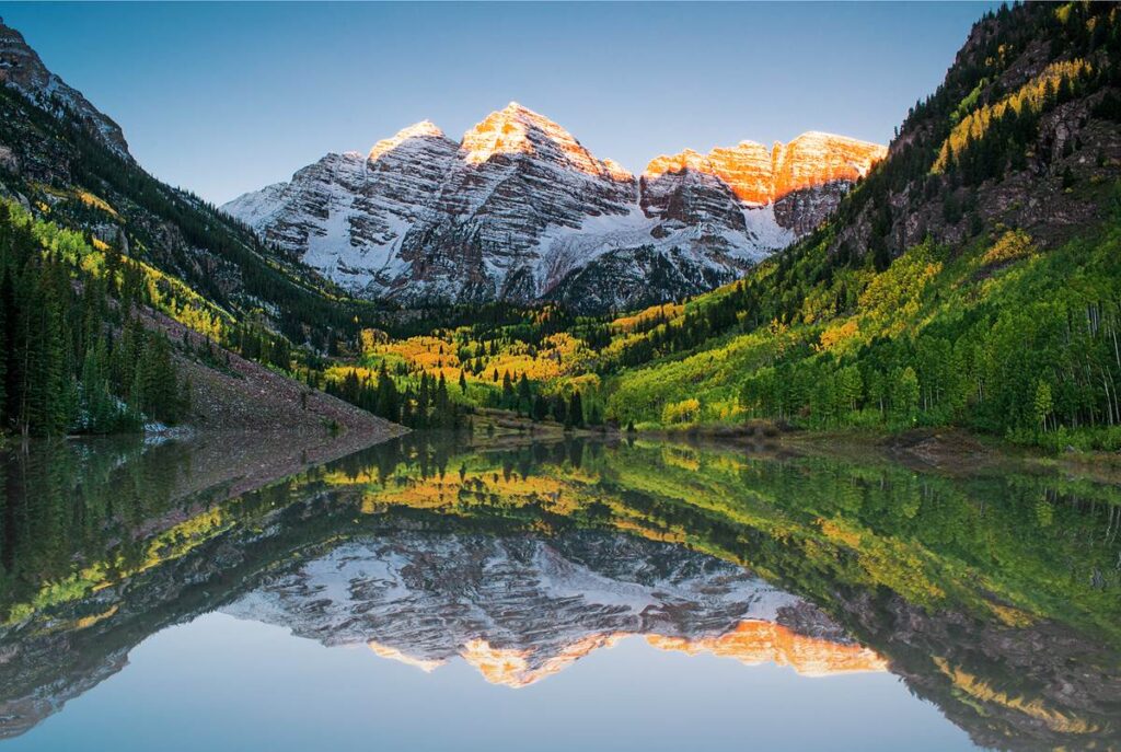 Snow-capped mountains at sunrise, reflected in a serene lake surrounded by lush green forest.