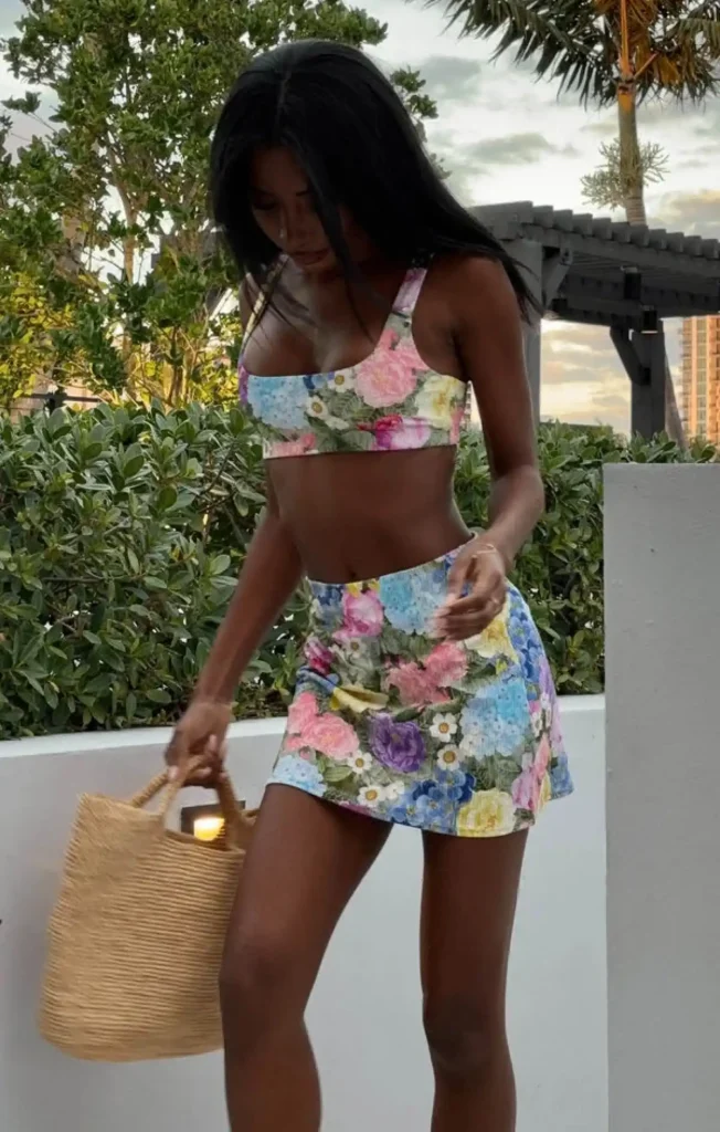 Model wearing a floral two-piece outfit, holding a woven bag, with greenery in the background. Perfect for summer fashion.