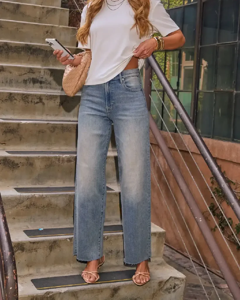 Woman in a white tee and wide-leg jeans standing on stairs, holding a phone and accessorized with a stylish bag.
