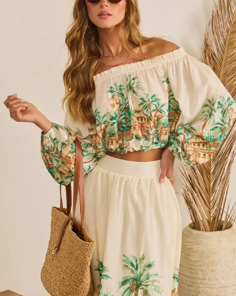 Stylish woman in an off-the-shoulder blouse and floral skirt, holding a woven bag, against a neutral backdrop.