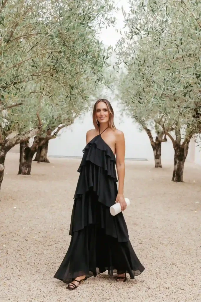 Woman in a flowing black tiered dress stands amidst olive trees, holding a white clutch, smiling at the camera.