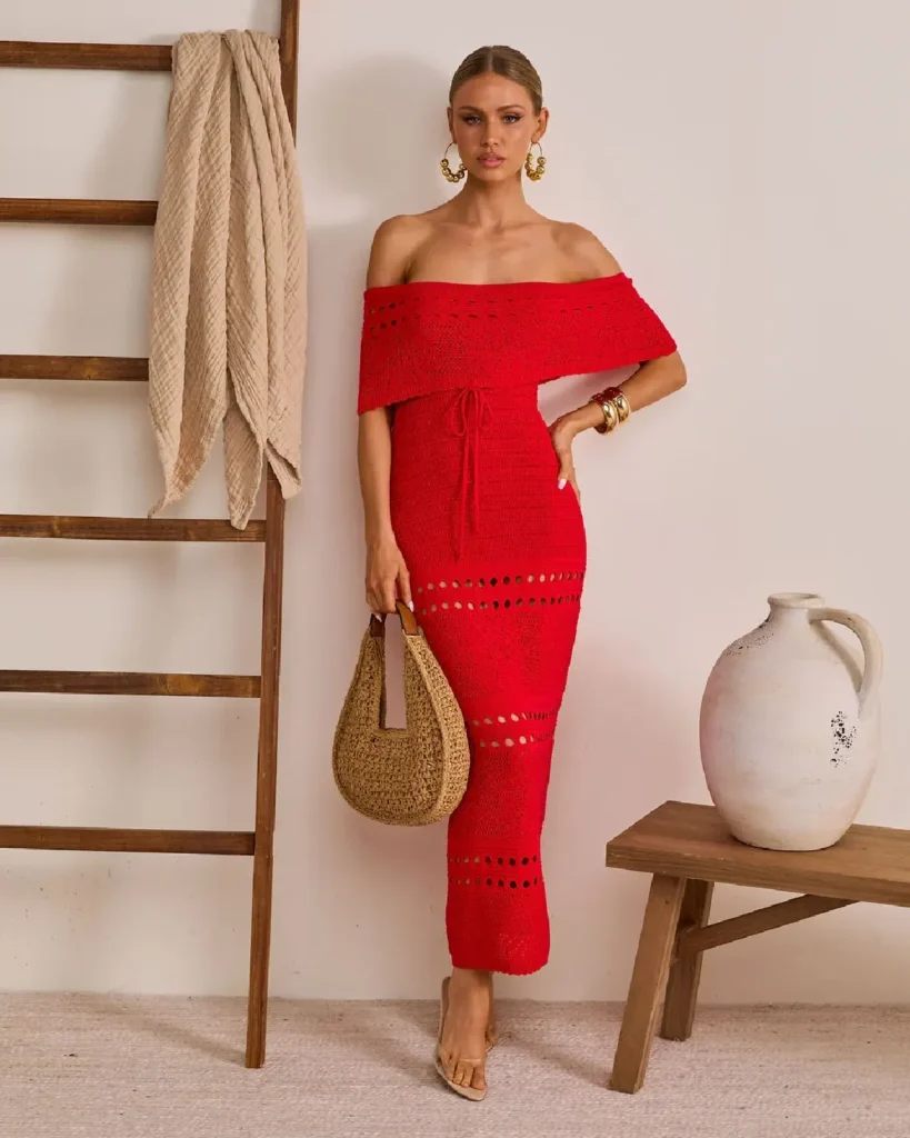 Stylish woman in a vibrant red off-shoulder dress, holding a straw bag, posed near a wooden ladder and ceramic vase.