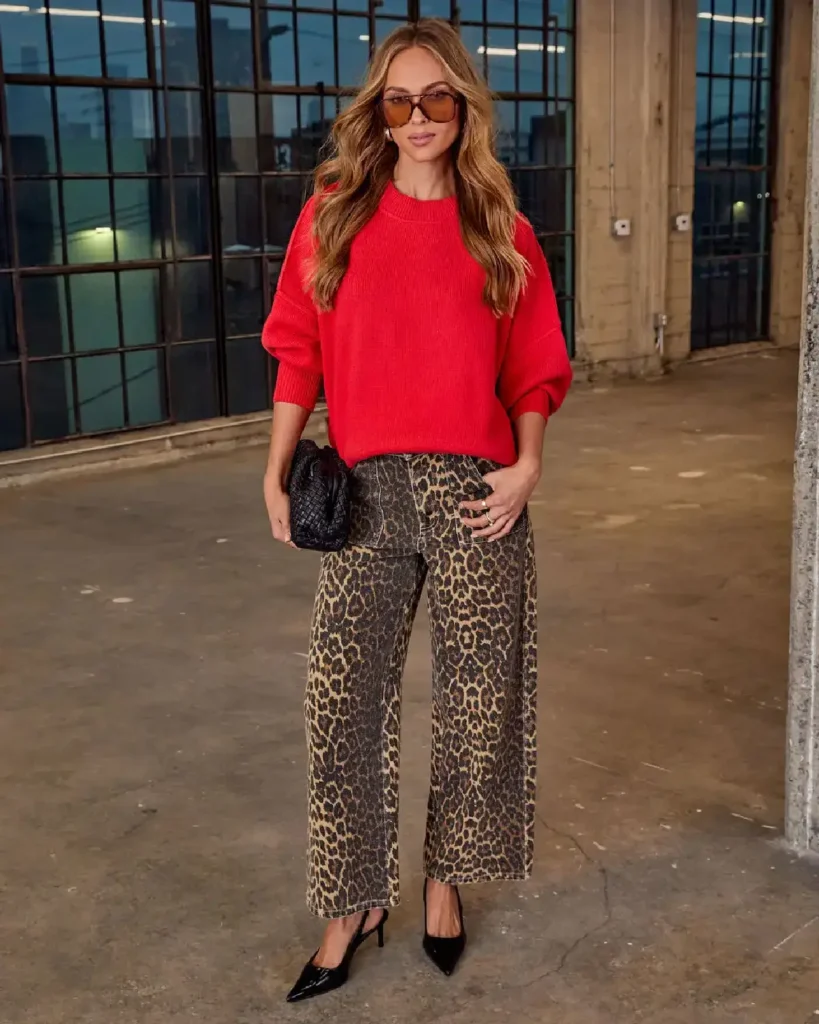 Stylish woman in a red sweater and leopard print pants poses indoors, showcasing a trendy fall outfit.