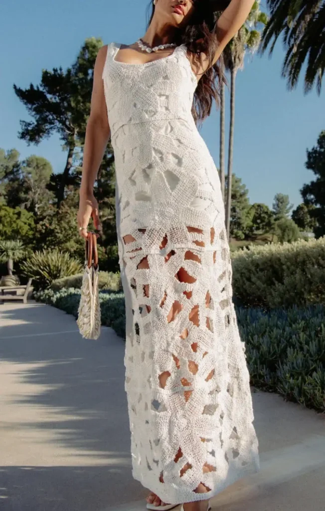 Model in a long white crocheted dress with cutouts, holding a purse, in a sunny outdoor setting with palm trees.