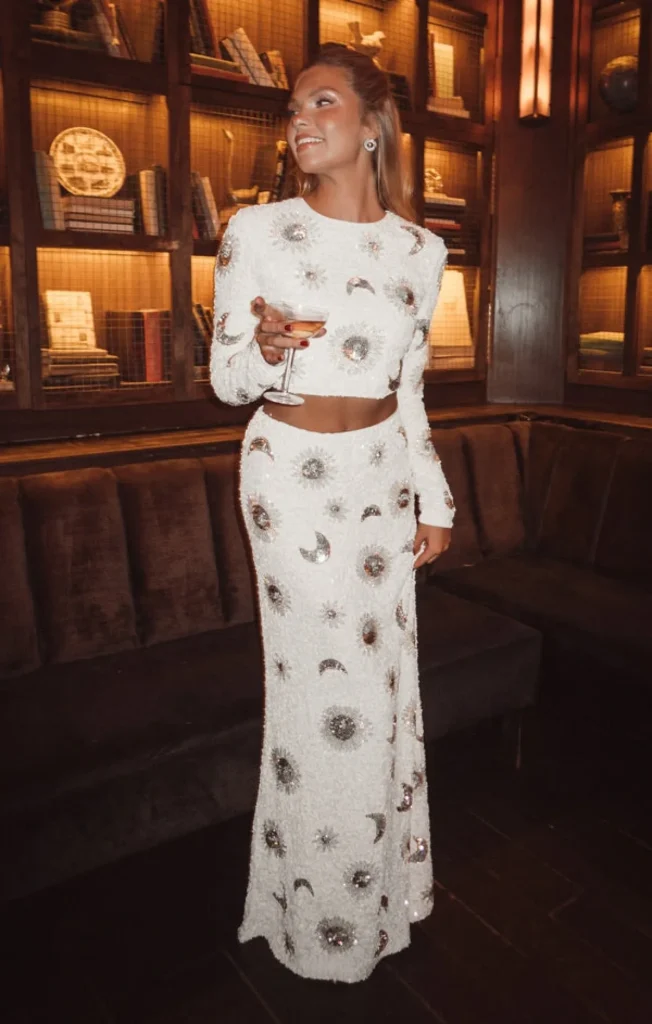 Elegant woman in a sparkling white two-piece outfit, holding a cocktail, against a stylish library backdrop.