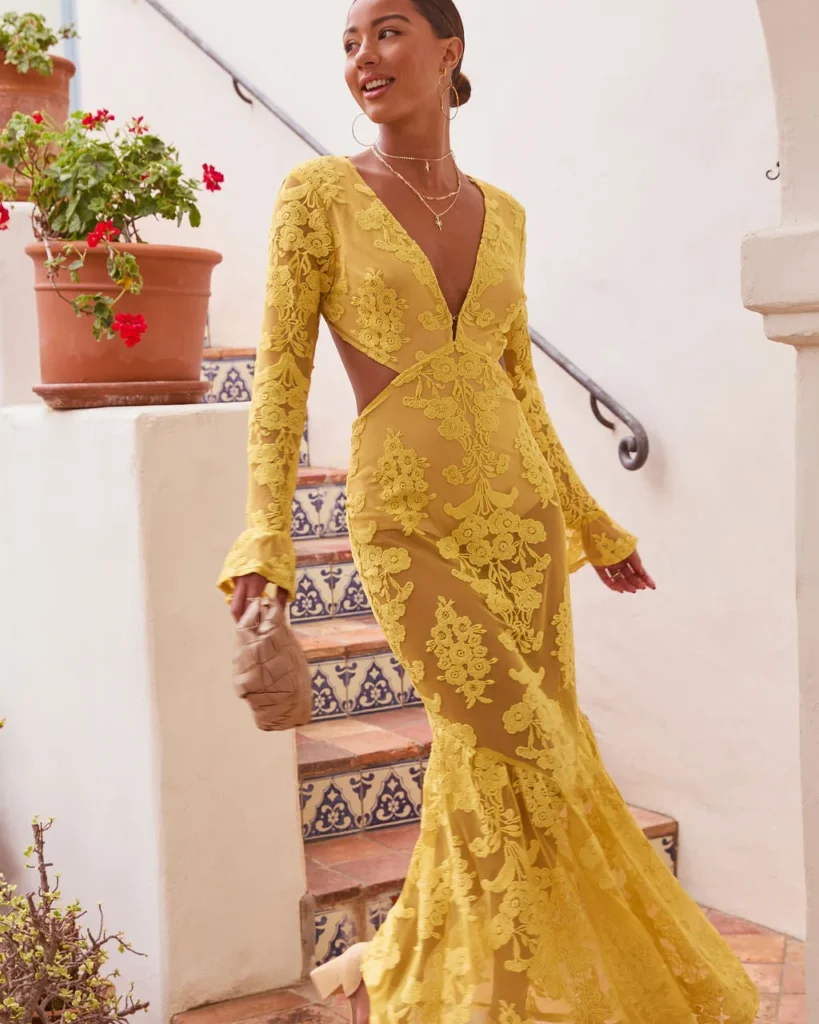 Woman wearing a vibrant yellow lace gown with floral designs, walking down a staircase surrounded by greenery.