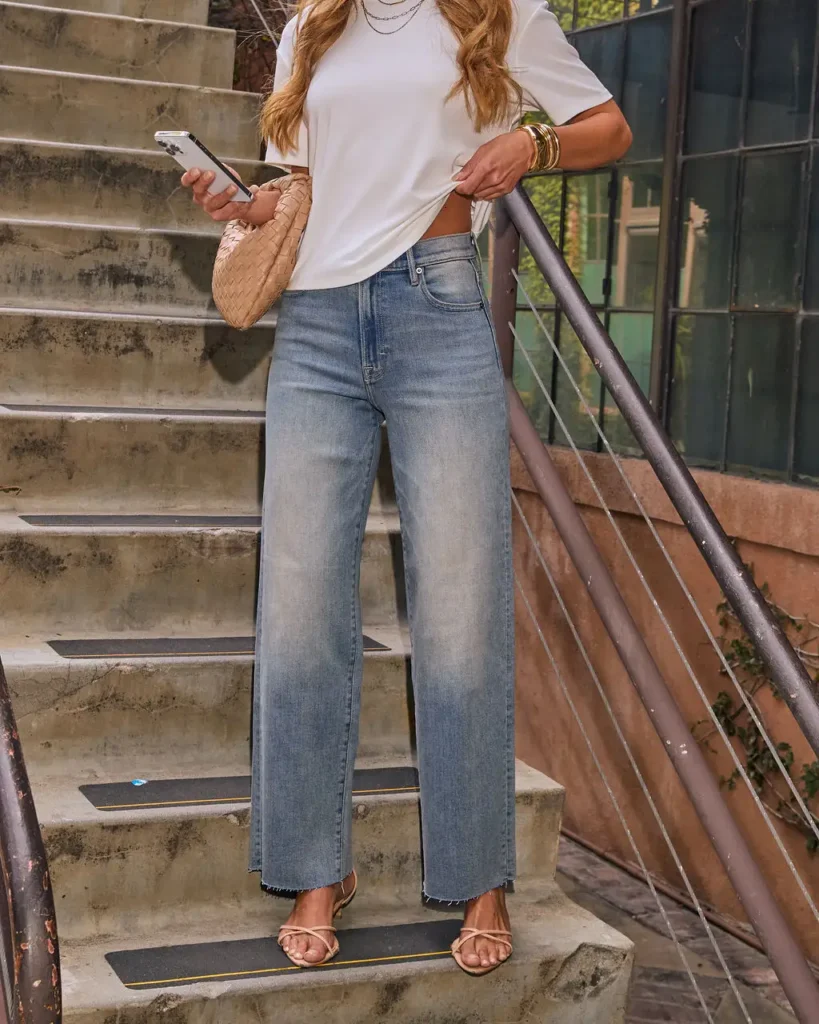 Fashionable woman on stairs, wearing a white shirt and wide-leg jeans, holding a phone and stylish bag.