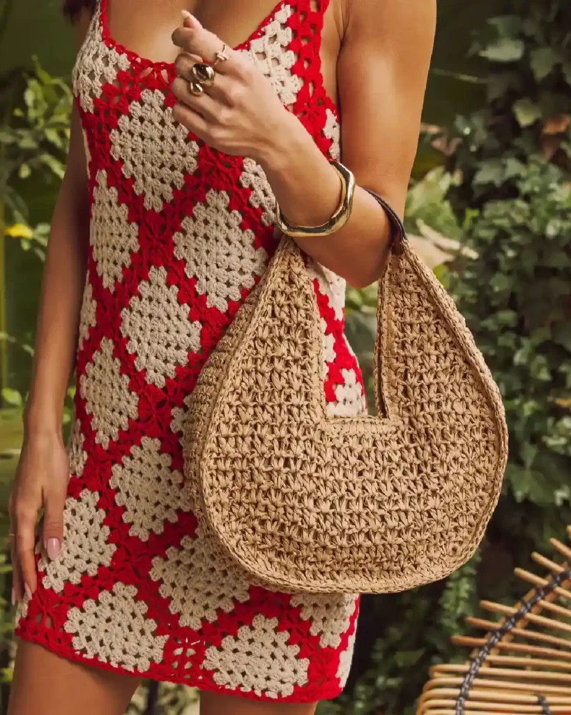 Woman in a stylish crochet dress holding a trendy woven handbag, set against a lush green backdrop.
