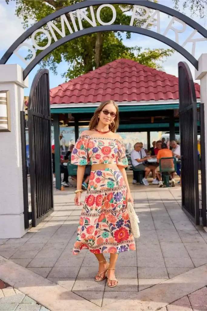 Woman in a colorful floral outfit poses at the entrance of Domino Park on a sunny day. Casual dining in the background.