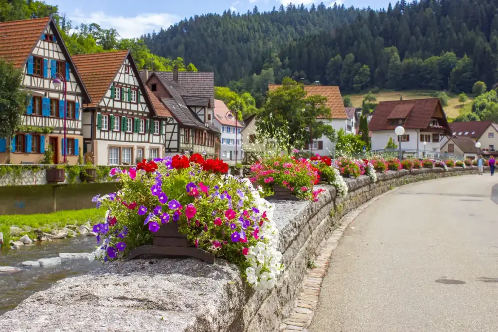 Charming village along a river, with beautiful flowers lining the stone path and picturesque houses against a mountain backdrop.