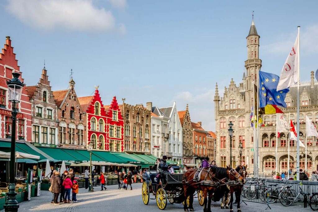 Colorful medieval buildings in Bruges city square, with flags and a horse-drawn carriage, under a blue sky.