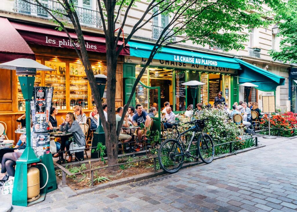 Outdoor Parisian café bustling with people, green awnings, and vibrant flowers on cobblestone street.