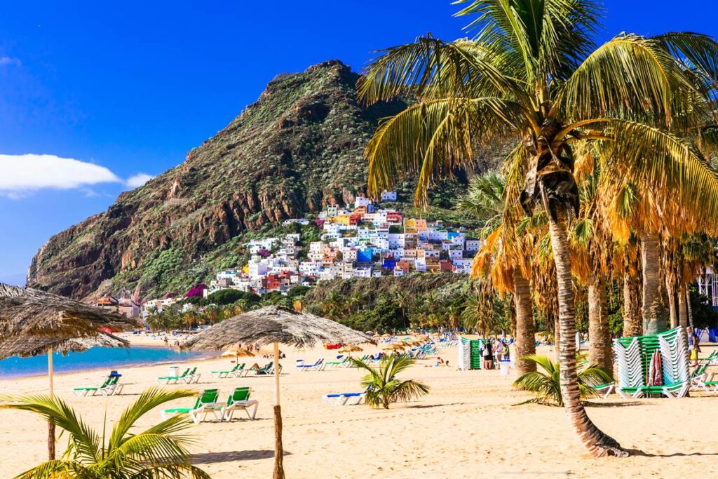 Colorful coastal village beside palm-lined sandy beach with sunbeds, nestled by a lush mountain under a clear blue sky.