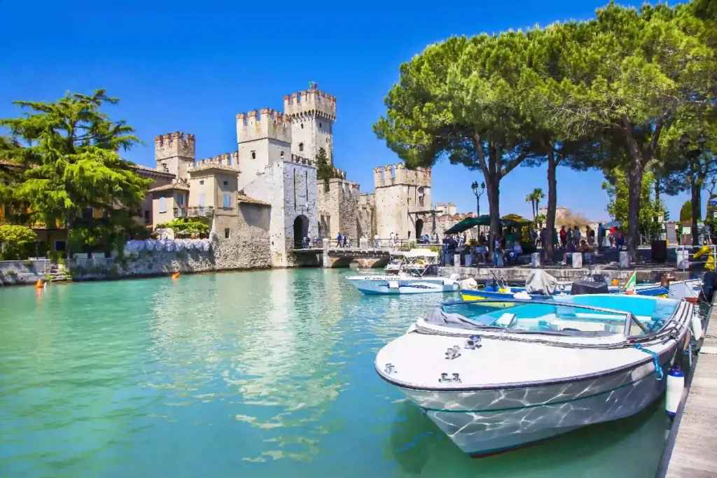 Scenic view of a medieval castle beside turquoise water, with boats and trees on a sunny day. Perfect for travel enthusiasts.