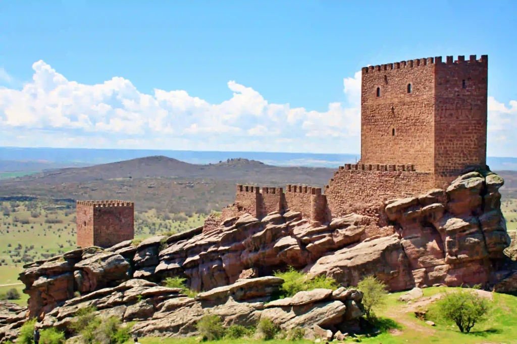Historical stone castle on rocky terrain against a blue sky with clouds, featuring lush green landscape in the background.