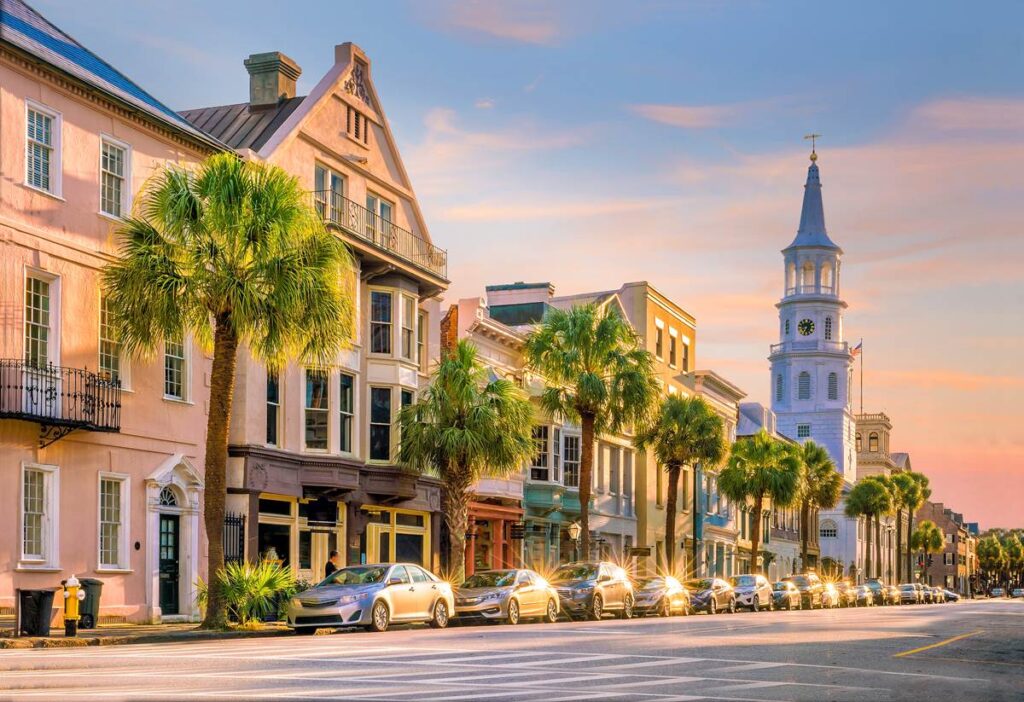 Historic Charleston street lined with colorful buildings, palm trees, and cars under a pastel sunset sky.