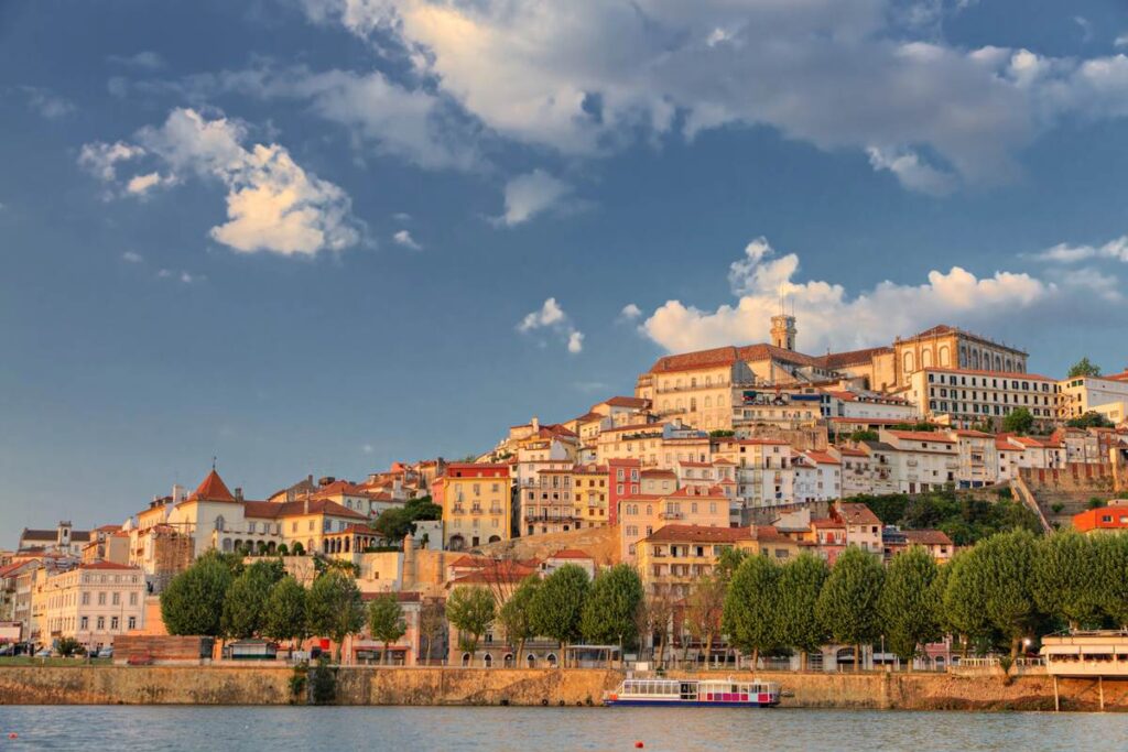 Coimbra skyline at sunset with historical buildings and trees by the river, under a partly cloudy sky.