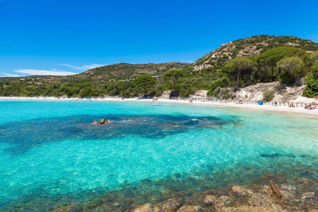 Turquoise water and sandy beach at Santa Giulia, Corsica, with lush greenery and mountains in the background.