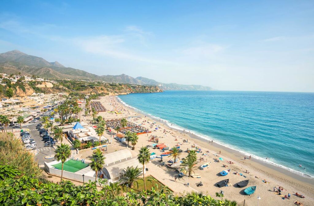 Sunny Mediterranean beach with vibrant umbrellas, azure sea, and distant mountains under a clear blue sky.