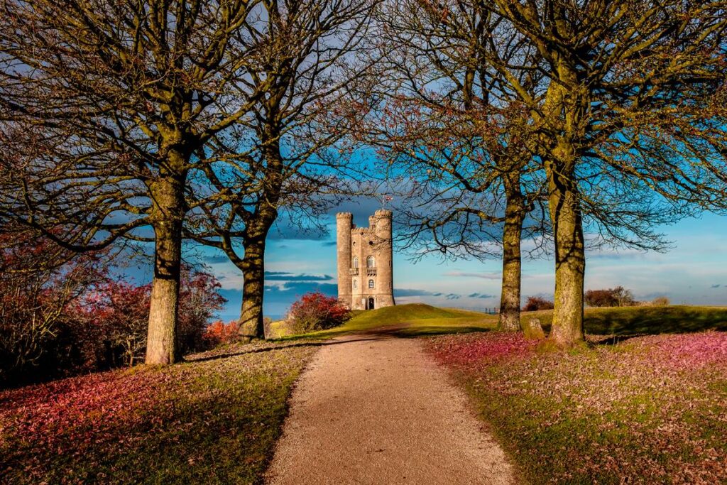 Pathway through trees leading to historic tower on a bright day with blue sky and lush scenery.