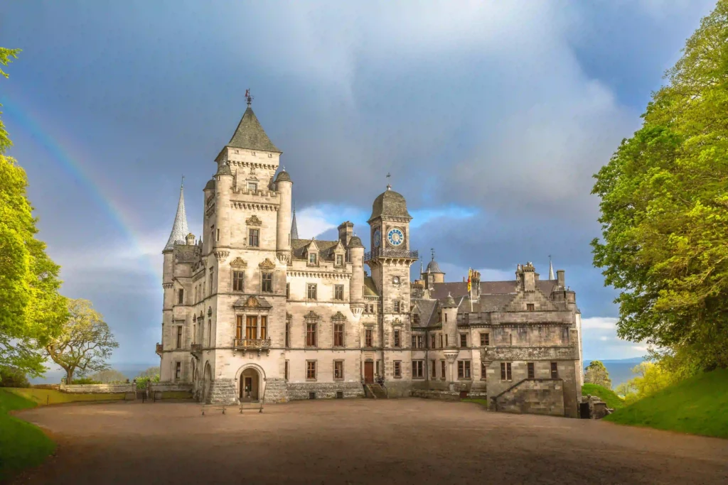 Beautiful historic castle with towers, clock, and lush greenery under a rainbow and dramatic sky.