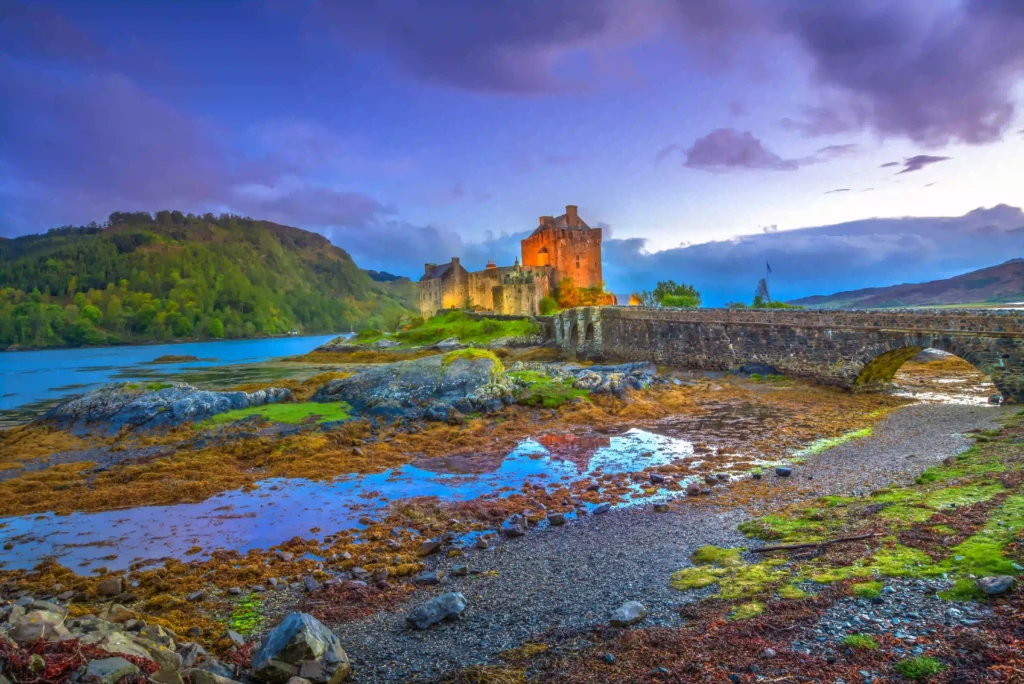 Scenic view of Eilean Donan Castle surrounded by water, hills, and vibrant reflections at sunset in Scotland.
