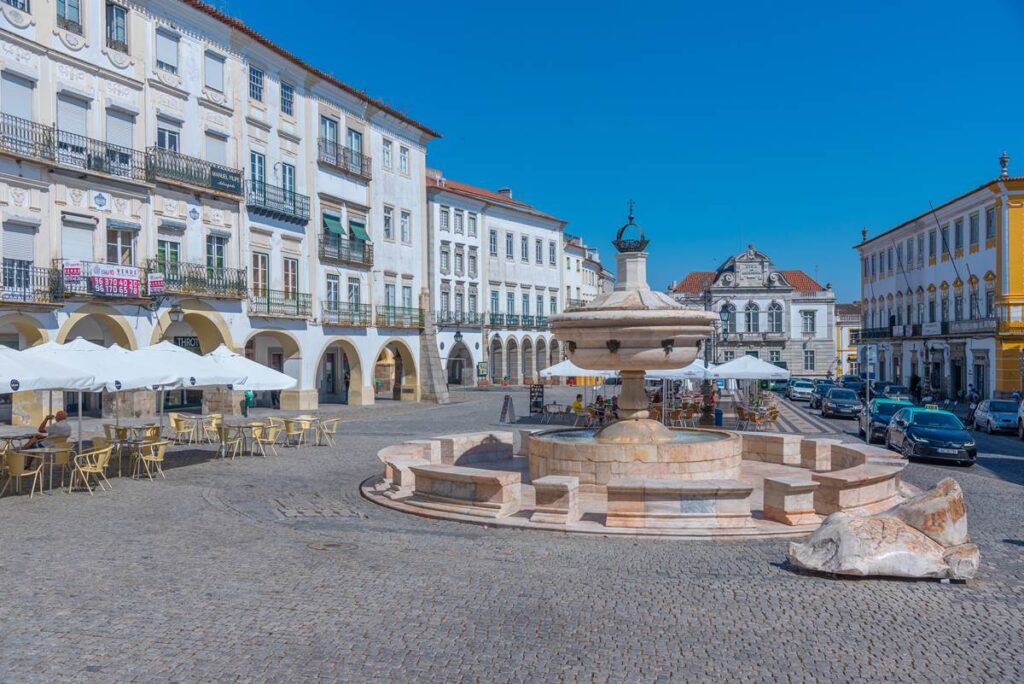 Historic town square with a stone fountain, outdoor cafes, and classic architecture under a clear blue sky.