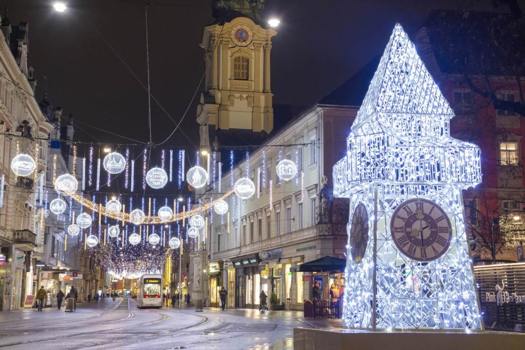 Festive city street with glowing clock tower and holiday lights illuminating a tram line, creating a magical winter scene.