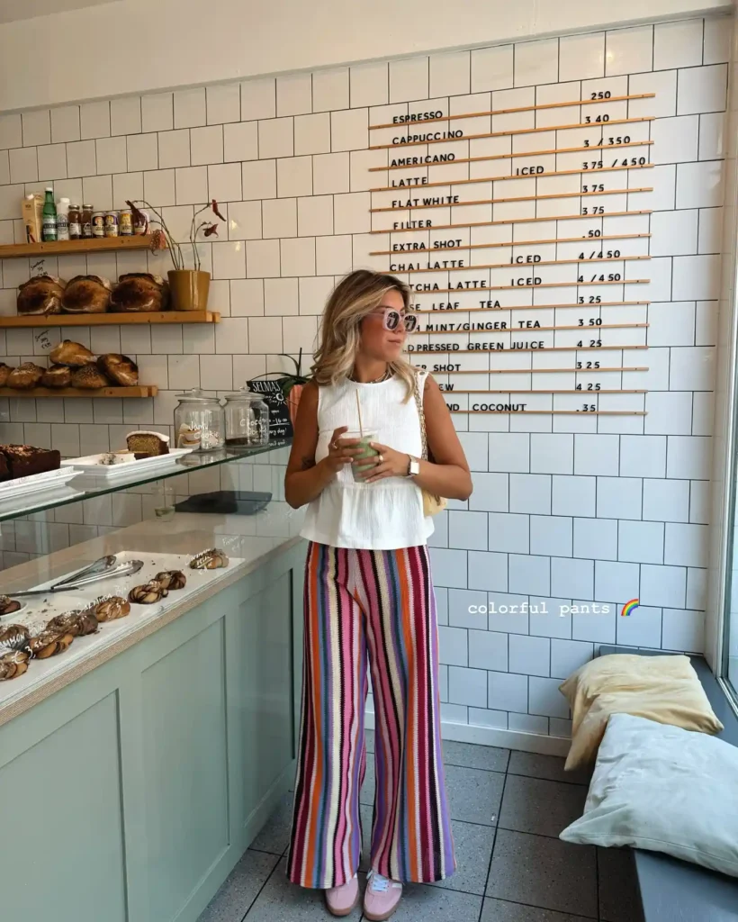 Woman in colorful striped pants standing in a stylish café with a menu on the wall and pastries displayed.