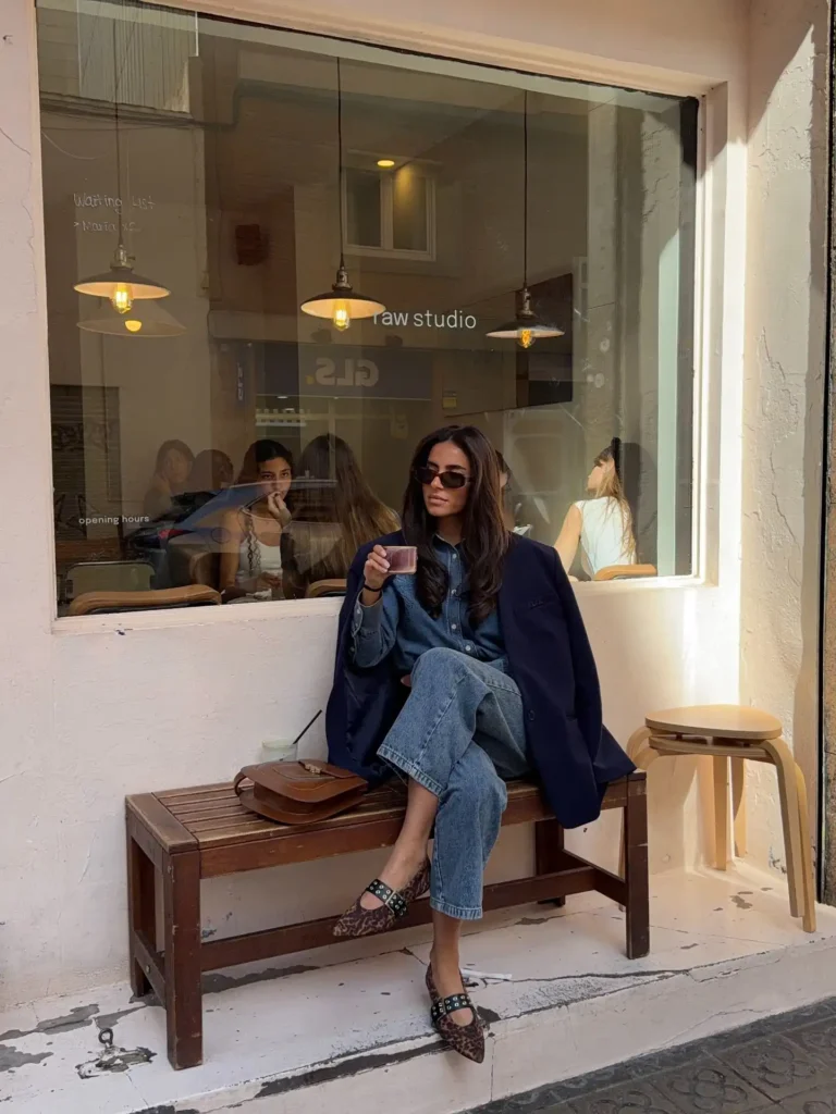 Fashionable woman sitting in a café, enjoying a drink, styled in denim and a chic jacket, with cozy indoor ambiance.