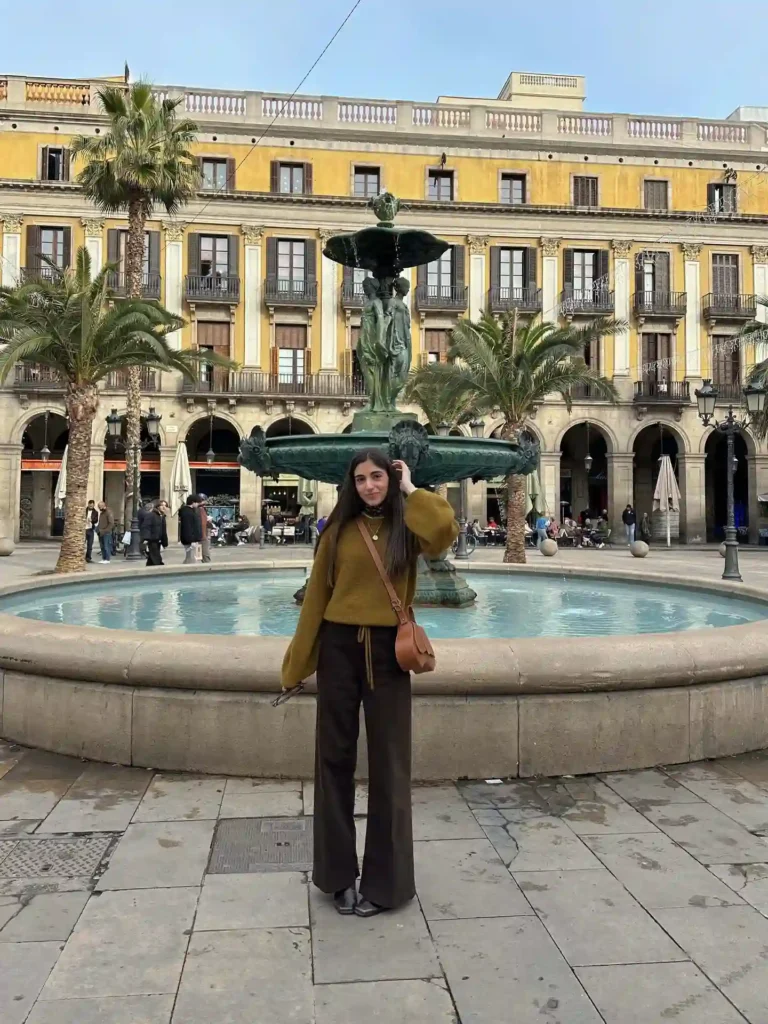 Person standing by a fountain in a square, surrounded by palm trees and historic buildings in a cityscape.