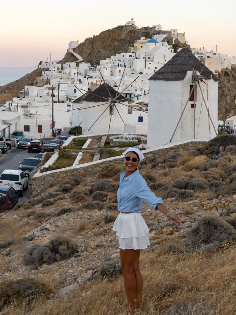 A joyful woman in a blue shirt and white skirt poses in front of Mykonos windmills and scenic white buildings at sunset.