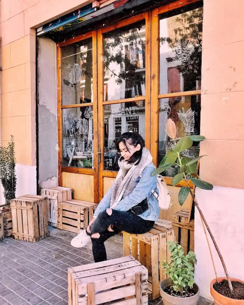Young woman in a cozy outfit sitting on wooden crates outside a trendy café, surrounded by plants and urban decor.