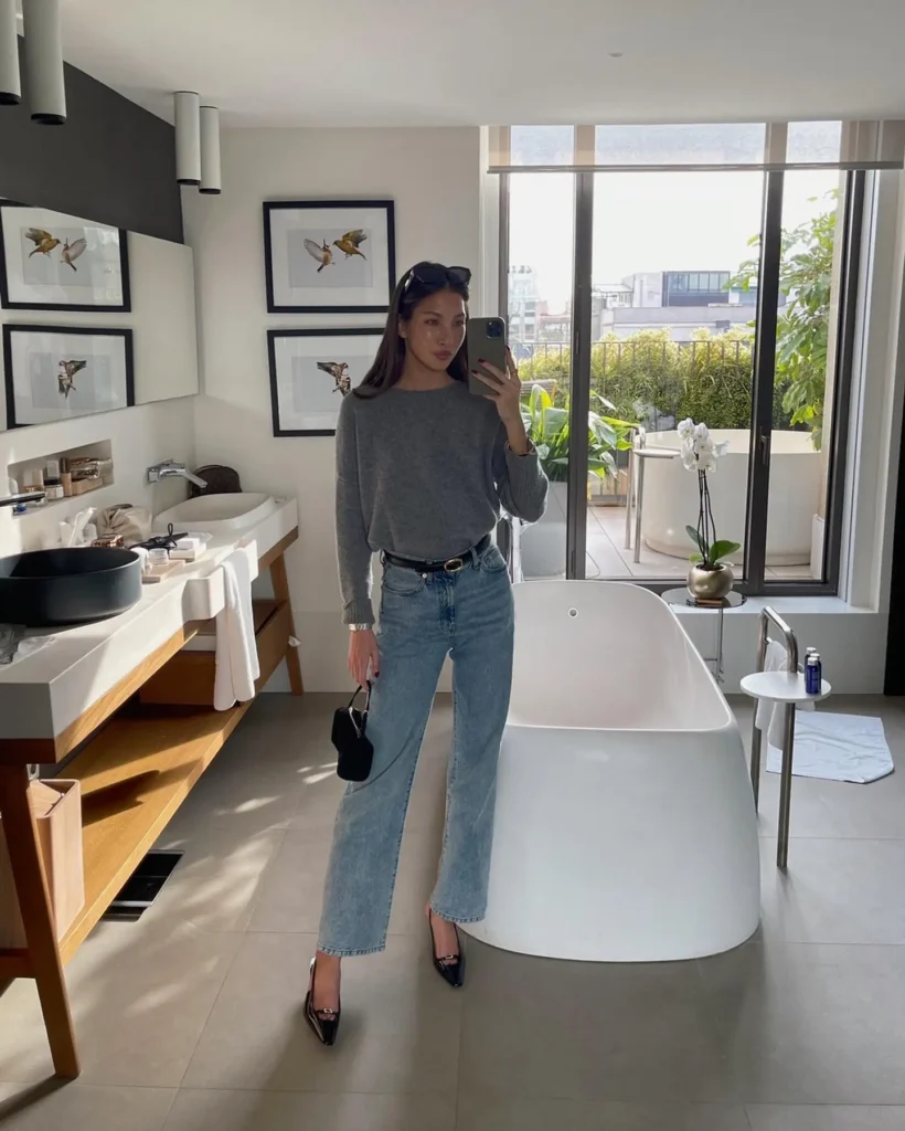 Woman in a gray sweater and jeans poses in a stylish bathroom with natural light and modern decor.