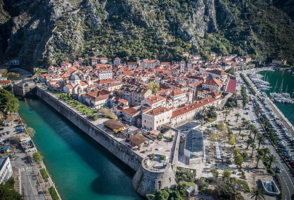 Aerial view of Kotor, Montenegro's old town with medieval architecture, city walls, and marina, surrounded by mountains.