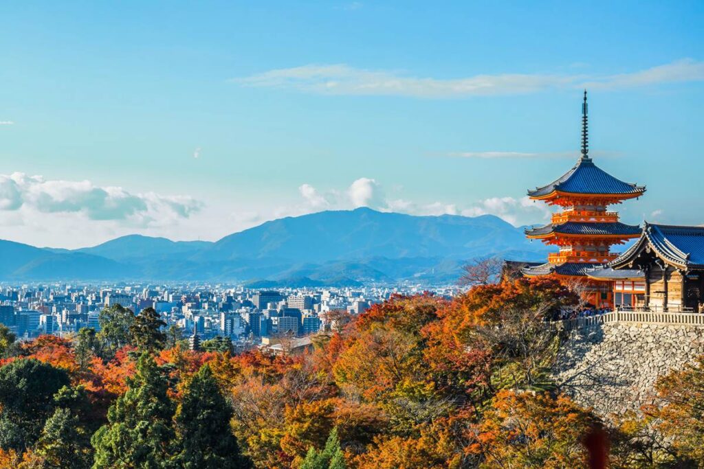 View of Kiyomizu-dera Temple with autumn leaves and Kyoto cityscape, Japan, against a backdrop of distant mountains.