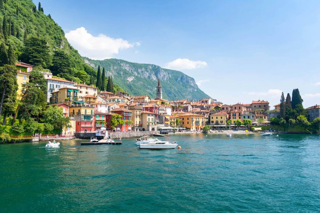Scenic view of colorful buildings by Lake Como with boats and lush green mountains in the background.