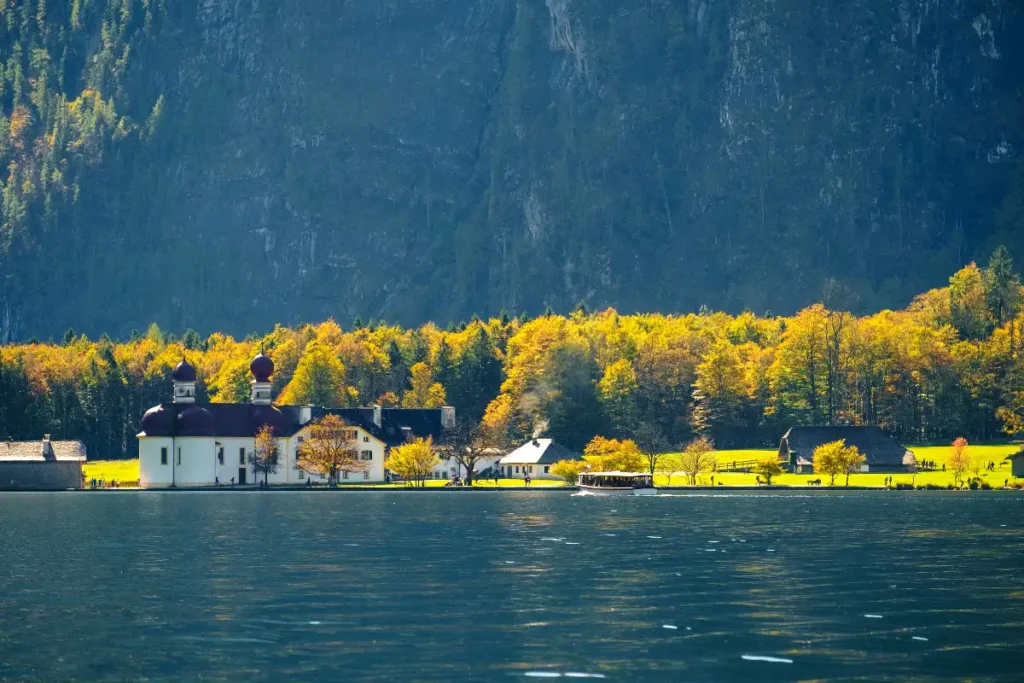 Scenic view of a serene lake surrounded by autumn foliage and charming countryside buildings in a mountainous landscape.