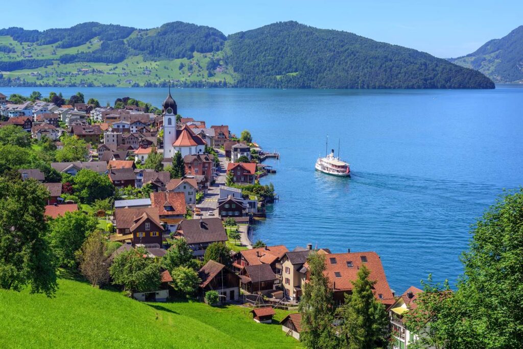 Scenic view of a lakeside village with a church, boat on lake, and green hills in the background under a clear blue sky.