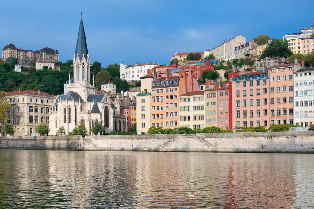 Colorful buildings and a historic church by the Saône River in Lyon, France, under a clear blue sky.