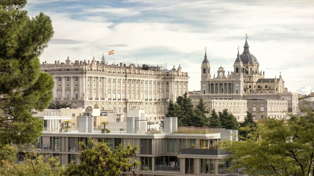 Royal Palace and Almudena Cathedral in Madrid, with trees and modern buildings, under a cloudy sky.