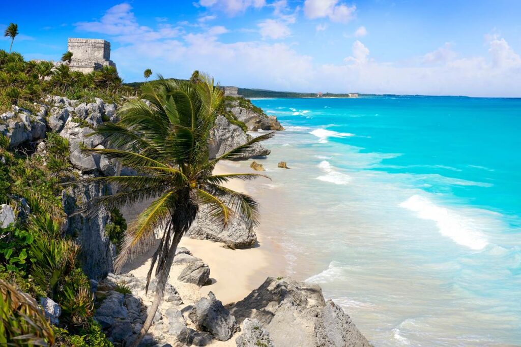 Stunning view of Tulum's beach with turquoise waters, rocky cliffs, and palm trees under a vibrant blue sky.