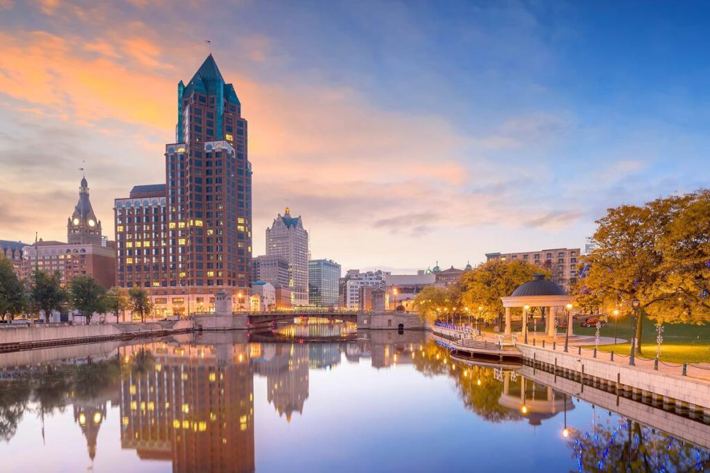 Skyline of Milwaukee at sunset, reflecting on the river with historic buildings and a gazebo.