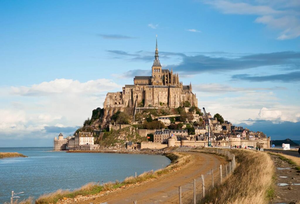 Mont Saint-Michel stands majestically against blue sky, surrounded by water and a winding path.