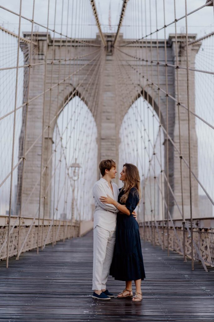 Couple embraces on Brooklyn Bridge, NYC, surrounded by cables and arches, creating a romantic atmosphere.