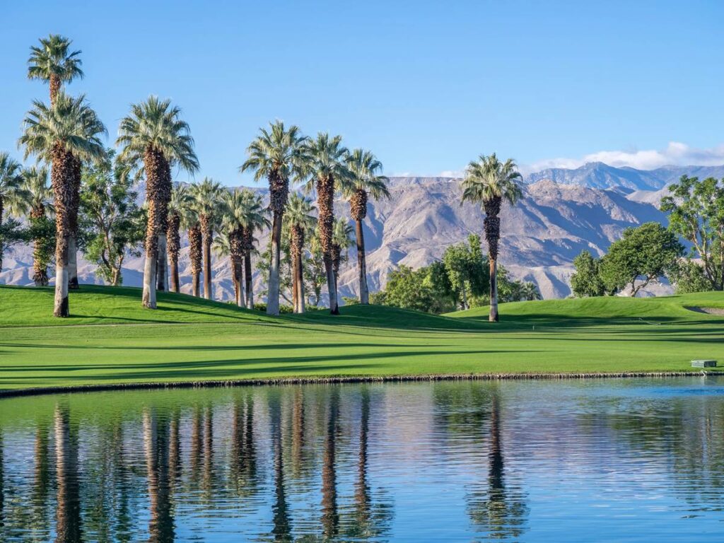 Lush green golf course with palm trees, scenic mountain backdrop, and reflecting pond under clear blue sky.